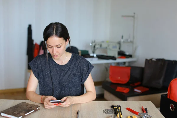 Artesana creativa sentada en el taller y usando un teléfono inteligente, artículos de cuero hechos a mano . — Foto de Stock