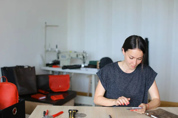Νέοι craftswoman κάθεται στο ατελιέ και χρησιμοποιώντας το smartphone, χειροποίητα δερμάτινα είδη. — Φωτογραφία Αρχείου