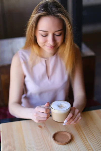 Piękna Pani siedzi w pobliżu drewnianym stole w przytulnej kawiarni z filiżanki cappuccino i pozowanie do zdjęć. — Zdjęcie stockowe