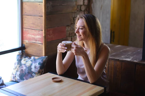 Młoda kobieta z filiżanki cappuccino w ręku siedział w pobliżu okna w przytulnej kawiarni. — Zdjęcie stockowe