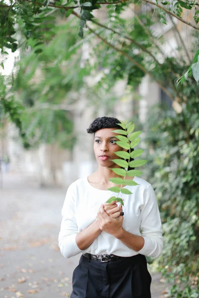 Menina preta bonita elegante de pé com folha verde e vestindo blusa branca e tendo franja . — Fotografia de Stock