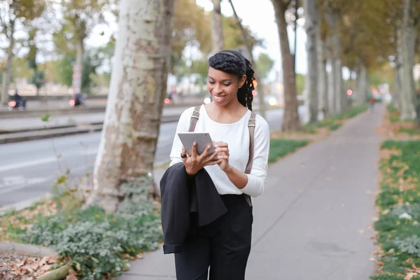 Black female student using tablet and walking on street with trees.