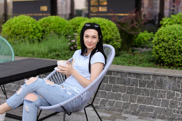Female person sitting in chair at cafe near green plants and drinking coffee.