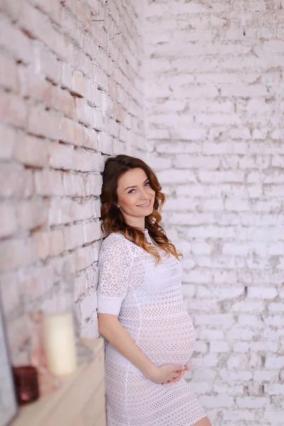 Mulher grávida vestindo vestido branco em pé no fundo da parede de tijolo e segurando barriga . — Fotografia de Stock