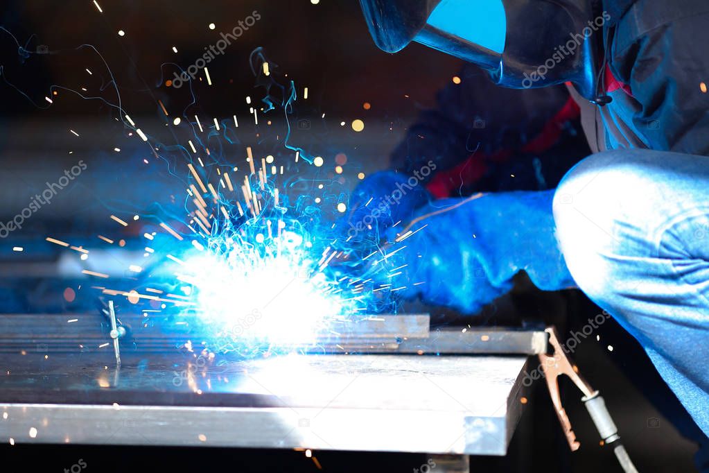 Male worker wearing construction gloves and mask welding metall construction, blue welding arc.