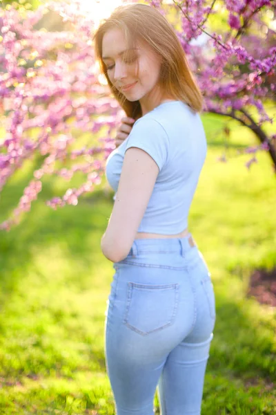 Charmig tjej klädd i jeans stående i solen strålar med blommande grenar bakgrund. — Stockfoto