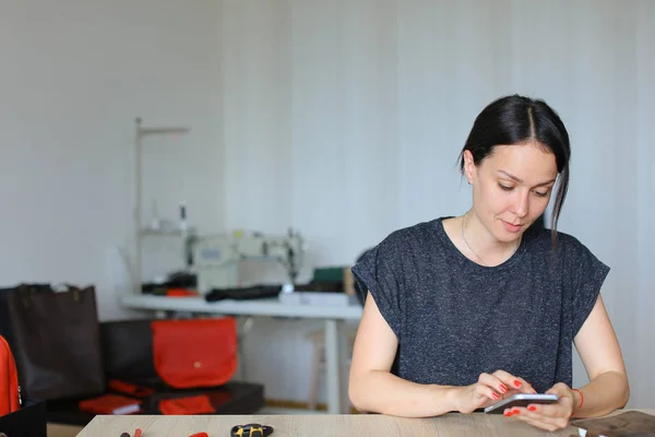 Artesana europea sentada en el taller y usando smartphone, artículos de cuero hechos a mano . — Foto de Stock