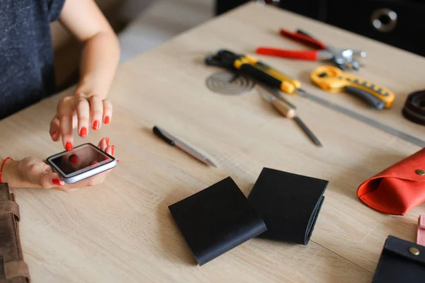 Female hands with red nails at leather atelier using smartphone, handmade wallets and tools on table.