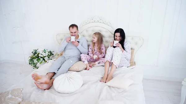 父母和小女儿躺在床上, 在卧室喝茶. — 图库照片