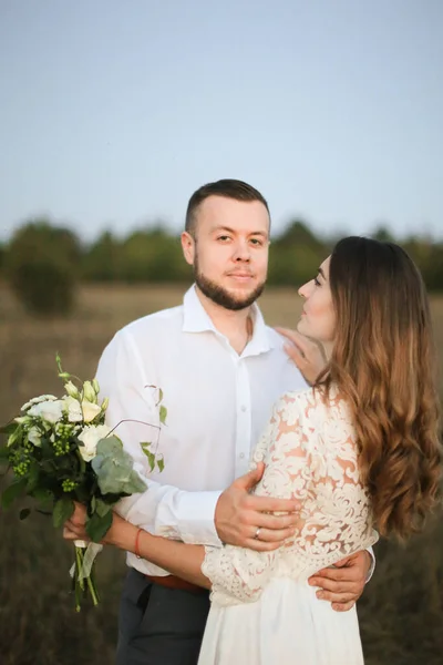 Yaklaş yakışıklı damat tarla arkasında bir buket çiçekle geline sarılıyor.. — Stok fotoğraf