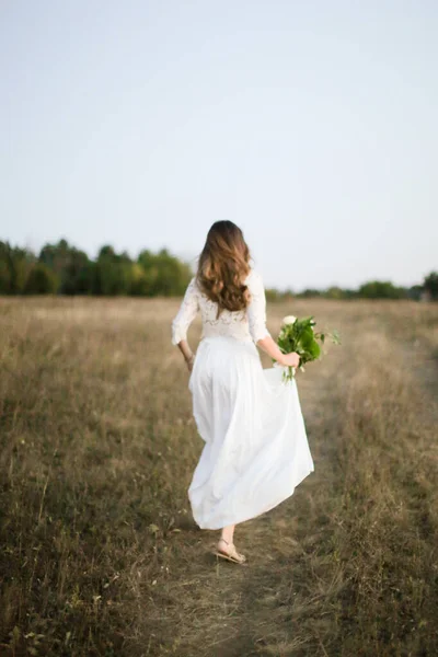 Europejska panna młoda w białej sukience spacery w polu z bukietem kwiatów. — Zdjęcie stockowe