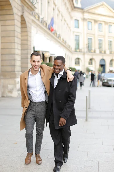 Caucasian smiling man in suit walking with afroamerican male person and hugging in city.