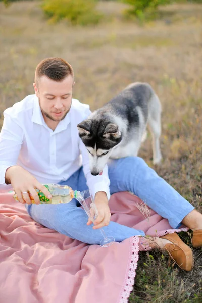 Young man pouring glass and sitting on pink plaid near husky outside. — Stock Photo, Image