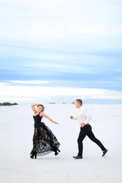 Jovem caucasiano mulher e homem correndo em estepe de inverno, vestindo vestido preto . — Fotografia de Stock