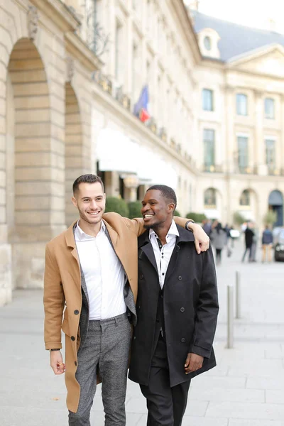Kaukasier glücklich Mann im Anzug zu Fuß mit afroamerikanischen männlichen Person und Umarmung in der Stadt. — Stockfoto