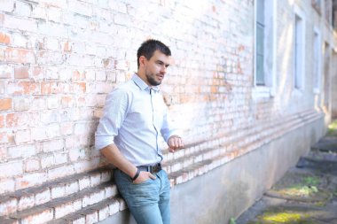 Genç yakışıklı adam duvarın yanında duruyor, önüne bakıyor ve pantolon ceplerinde el ele tutuşuyor..