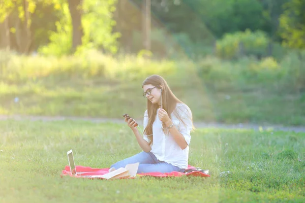 Niña sentada en el parque, sonriendo y trabajando en su portátil, escuchando música — Foto de Stock