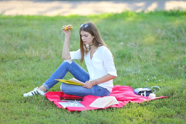 Giovane ragazza seduta sull'erba nel parco e lavora in un computer portatile e mangiare fast food — Foto Stock