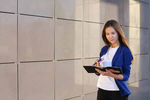 Joven adulto niña estudiante sostiene en las manos diario, cuaderno, Los estudiantes trabajan, tarea y escribe pluma contra el telón de fondo del centro de negocios . — Foto de Stock