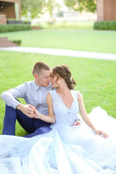 Caucásico bonita novia sentado con el novio y besándose en la hierba en el parque, usando vestido blanco . — Foto de Stock