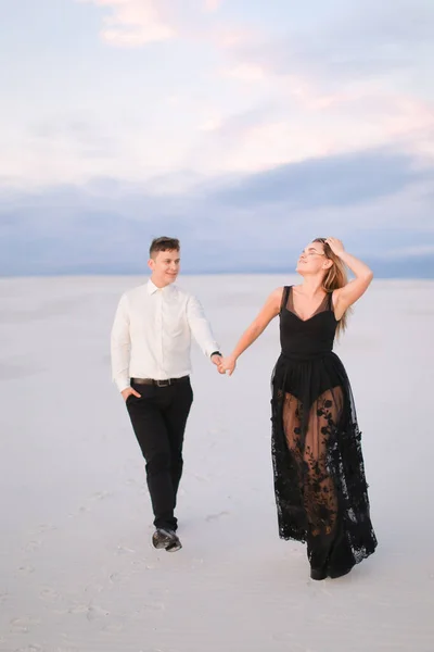 Man and blonde woman wearing black dress walking in snowed steppe and holding hands. — Stock Photo, Image