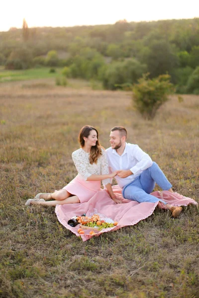 Gelukkig paar zitten op roze ruit met fles en fruit in het veld. — Stockfoto