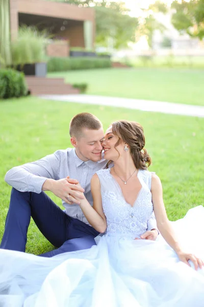 Caucasian nice bride sitting with groom and kissing on grass in park, wearing white dress. — Stock Photo, Image