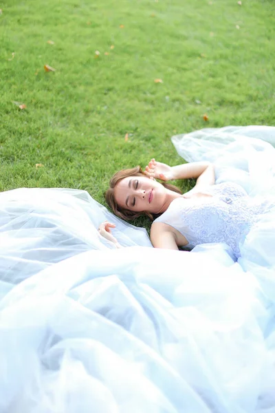 Innocent pretty bride lying on grass in oark and wearing white dress.