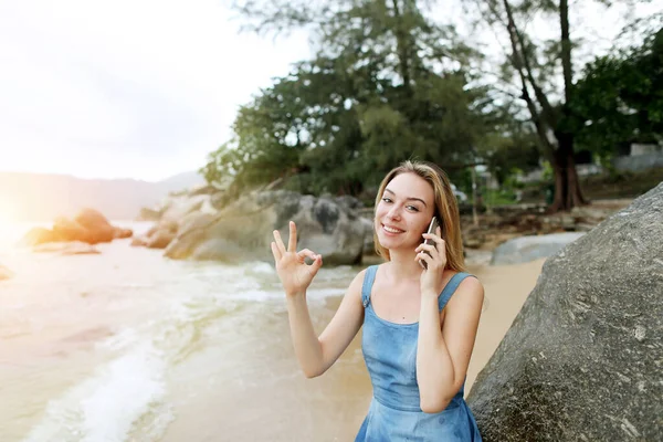 Foto de luz soleada de la joven mostrando ok y sentado en la orilla del mar, hablando por teléfono inteligente Hawaii . — Foto de Stock