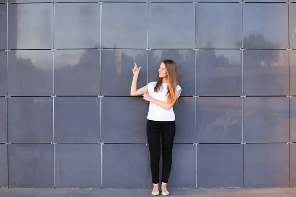 Jeune fille énergique femelle levé les bras et les points à l'espace publicitaire, vaut exactement en pleine croissance sur fond de mur du centre d'affaires . — Photo