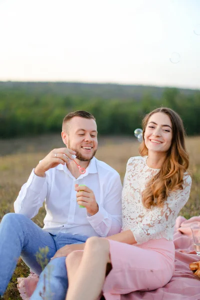 Junger lächelnder Mann bläst Seifenblasen in der Nähe kaukasischer Mädchen und sitzt auf Plaid in der Steppe. — Stockfoto