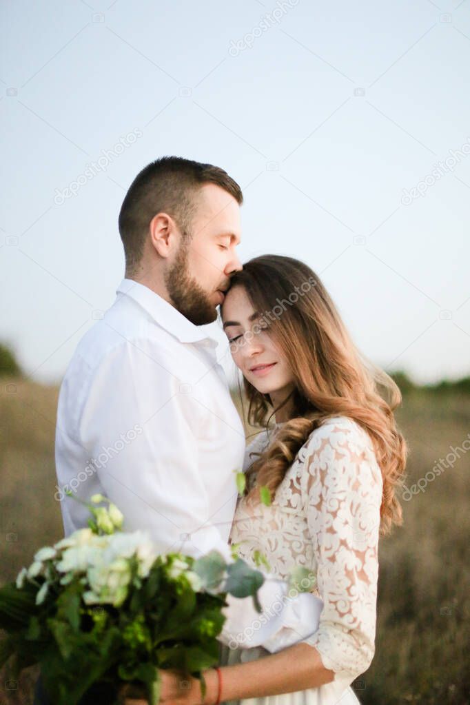 Close up groom hugging bride with bouquet of flowers in field background.