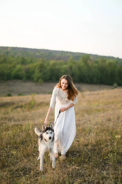 Caucasian beautiful bride walking in steppe with dog and wearing white dress. — Stock Photo, Image