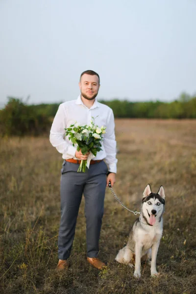 Fiatal vőlegény áll csokor fehér virágok sztyeppén közel husky. — Stock Fotó