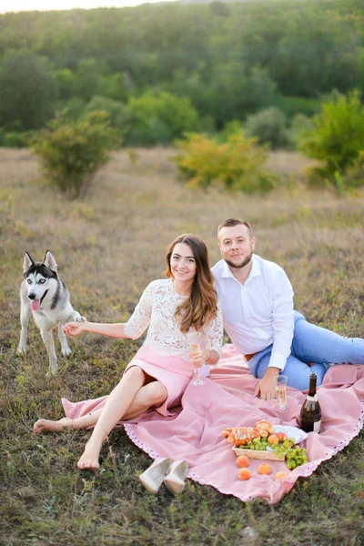 Young woman and man sitting on pink plaid in steppe near dog husky. — Stock Photo, Image