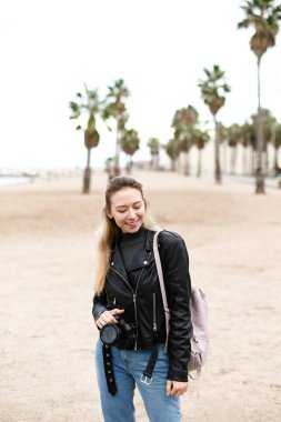 Young woman walking on beach with camera in leather jacket.