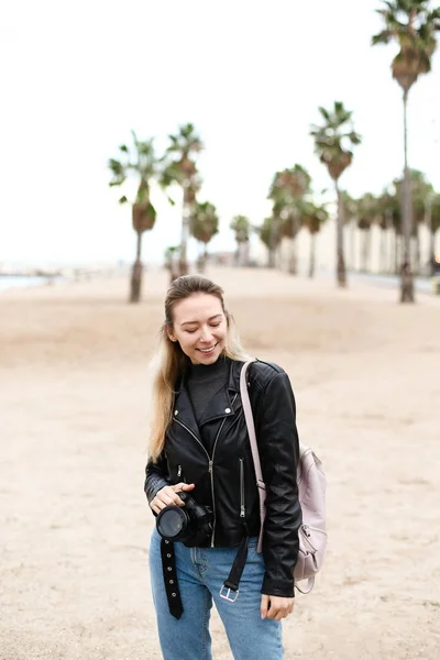 Jovem mulher andando na praia com câmera em jaqueta de couro . — Fotografia de Stock