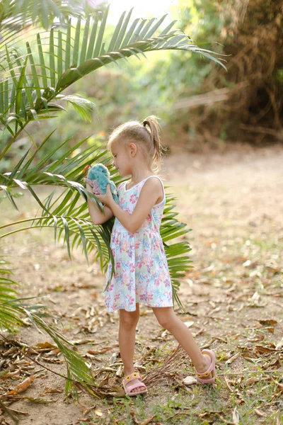 Niña jugando con juguete y de pie cerca de hojas de palma . — Foto de Stock