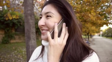 Slow Motion Asyalı mutlu kız beyaz kazak giyiyor, akıllı telefonla konuşuyor ve parkta yürüyor..