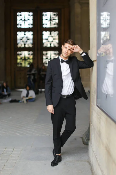 Junger gutaussehender Mann steht und lehnt am Gebäude, trägt schwarzen Anzug und Fliege. — Stockfoto