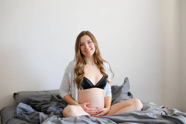 Schwangere Frau in schwarzen Dessous hält ihren Bauch und sitzt auf dem Bett. — Stockfoto