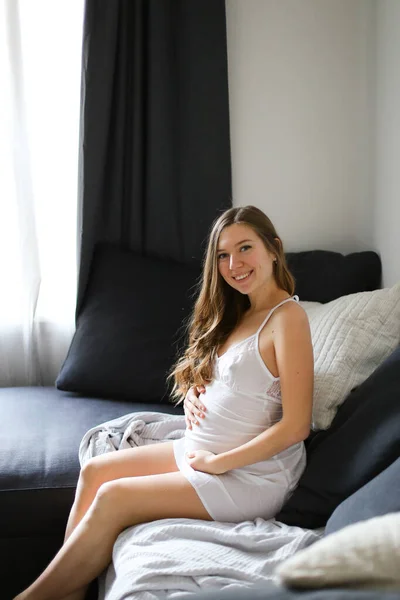 Молода біла вагітна жінка сидить на ліжку в білій сукні . — стокове фото