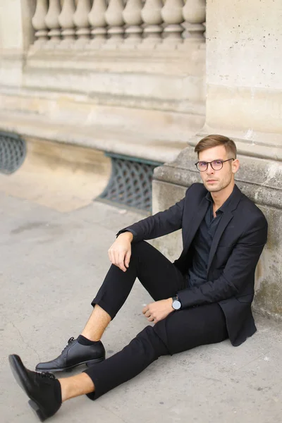 Joven europeo sentado en el suelo de la acera y vestido con traje negro . — Foto de Stock
