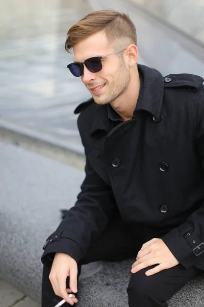 Jeune homme blond souriant portant une veste noire assis sur la pyramide du Louvre et fumant . — Photo