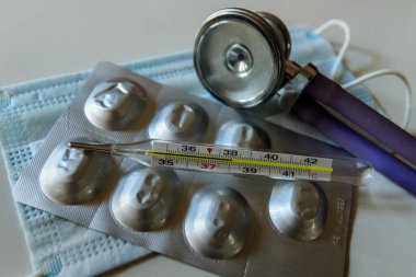 Cıva termometresi maskenin arka planında yüksek sıcaklık olduğunu gösteriyor. Su toplayan kutuda haplar ve stetoskop. Coronavirus hastalığı 2019 (Covid-19).