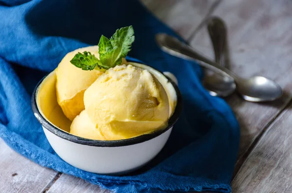 Gelato rinfrescante al mango / frutto della passione in una tazza di ferro su un tavolo di legno chiaro con tessuti blu e cucchiai sullo sfondo . — Foto Stock