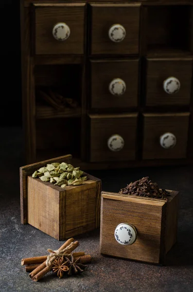 Spices in wooden boxes,chest of drawers in the background. Clove, anise, cinnamon, cardamom. Dark concrete background, vertical, copy space
