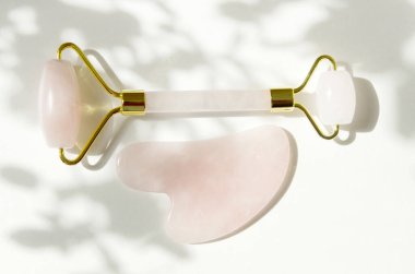 Pembe Gua Sha masaj aleti. Rose Quartz silindiri. Beyaz arka plandaki yaprakların gri gölgesi. Evde yüz bakımı, yaşlanmayı önleme ve kaldırma terapisi. - Görüntü