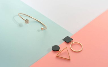 Kalp şeklinde altın bilezik ve geometrik modern küpeler pastel renkli arka planda.