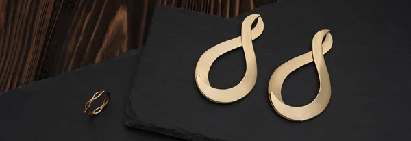 Szeroki strzał w kształcie złotego symbolu nieskończoności kolczyki i pierścień na ciemnej kamiennej powierzchni — Zdjęcie stockowe